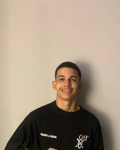 Retrato de Daniel de Lima Melo, sorrindo, de camiseta preta, diante de uma parede clara.